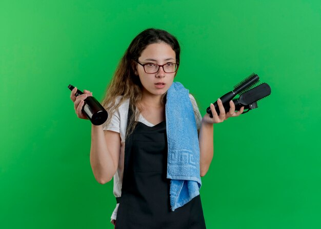 Young professional hairdresser woman in apron with towel on shoulder holding hair brushes and spray looking aside with serious face 