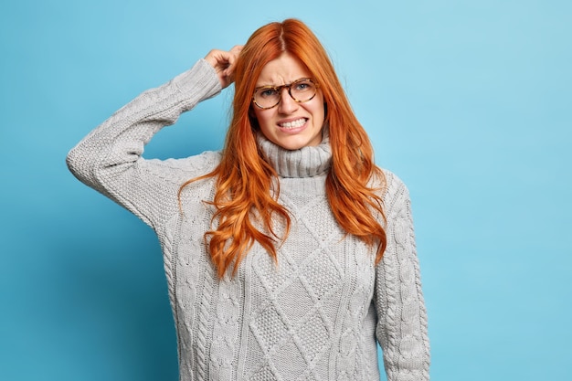 Young puzzled ginger woman scratches head clenches teeth and looks with dissatisfaction tries to solve problem or remember something important has bad memory.