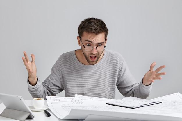 Young puzzled young architect feels indignant, looks ar drawings on table, realizes that he made mistake