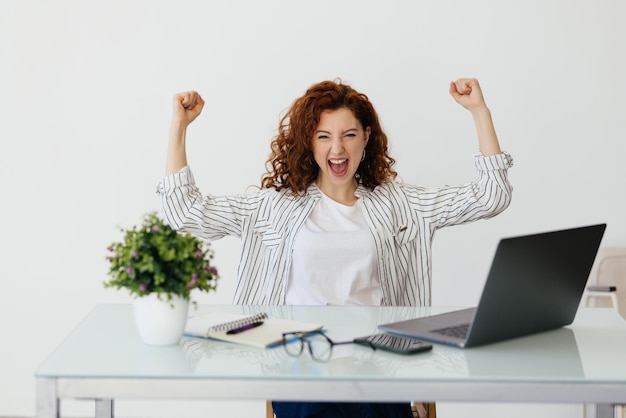 Free photo young redhead curly woman working with her laptop raising fist after a victory winner concept