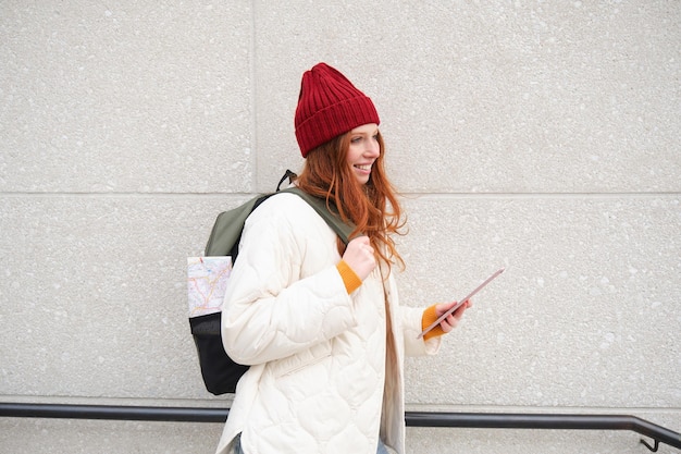 Free Photo young redhead girl walks with digital tablet around city stands on street with backpack and gadget l