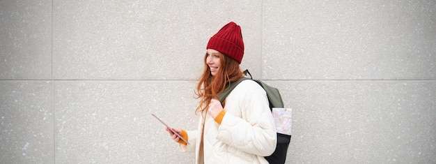 Free Photo young redhead girl walks with digital tablet around city stands on street with backpack and gadget