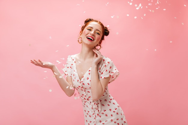 Young redhead lady in white dress smiles coquettishly. Woman with yellow eye shadows posing on pink background with confetti.