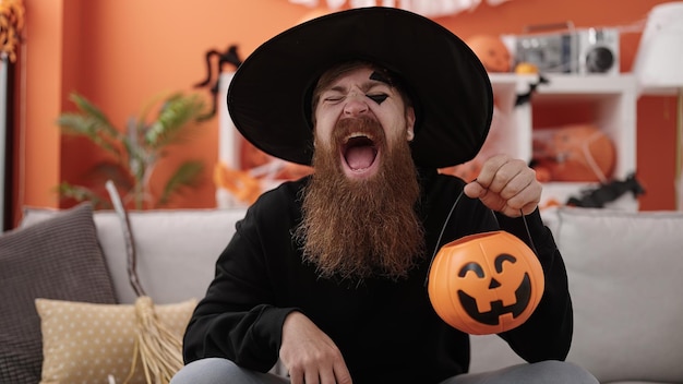 Young redhead man wearing wizard costume having halloween pumpkin basket at home