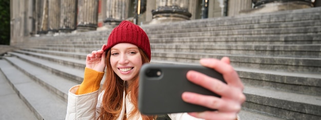 Free photo young redhead tourist takes selfie in front of museum on stairs holds smartphone and looks at mobile