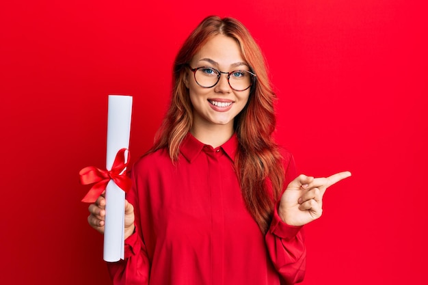 Young redhead woman holding graduate degree diploma smiling happy pointing with hand and finger to the side