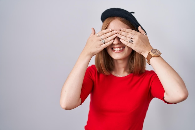 Free photo young redhead woman standing wearing glasses and beret covering eyes with hands smiling cheerful and funny blind concept