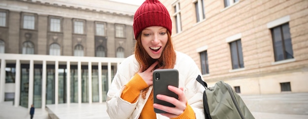 Free photo young redhead woman with smartphone sitting outdoors with backpack student looking at her mobile