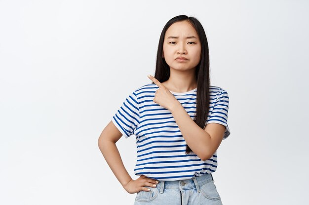 Young reluctant asian woman pointing finger at upper left corner grimacing disappointed being unsatisfied with smth standing over white background