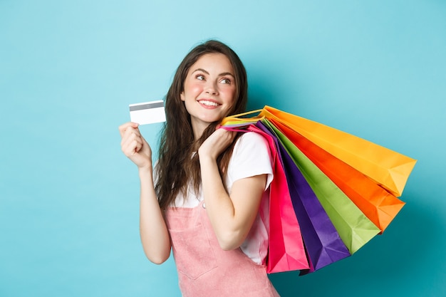 Young satisfied woman smiling, showing plastic credit card and holding shopping bags, buying with contactless payment, standing over blue background