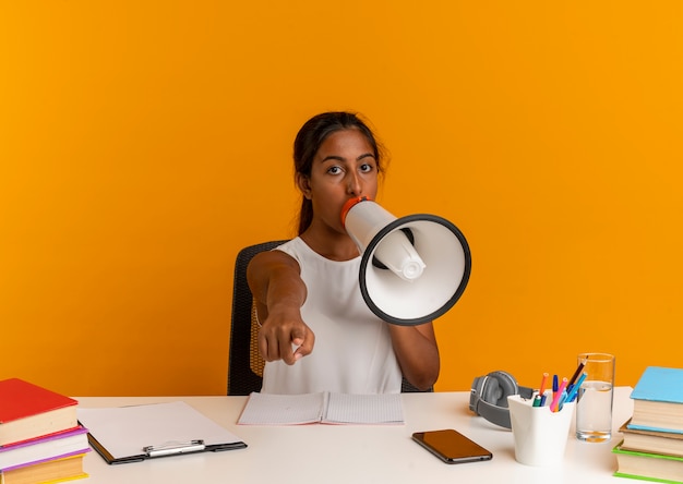 young schoolgirl sitting at desk with school tools speaks on loudspeaker 