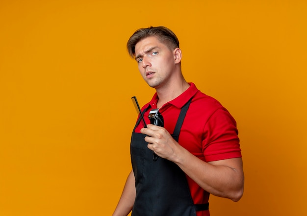 Young serious blonde male barber in uniform holds comb and hair clipper isolated on orange space with copy space