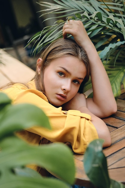 Young serious brown haired woman in yellow shirt lying on hand thoughtfully looking in camera with green leaves around in city park