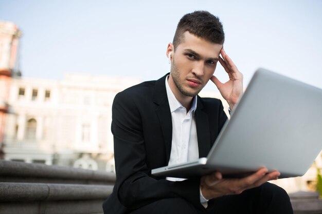 Young serious businessman in classic black jacket and white shirt with wireless earphones thoughtfully looking in camera with laptop in hand outdoor