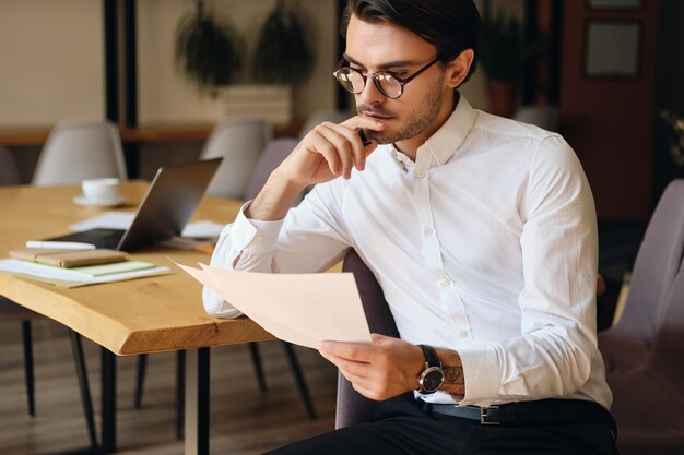 Young serious businessman in eyeglasses thoughtfully reading papers while working in modern office