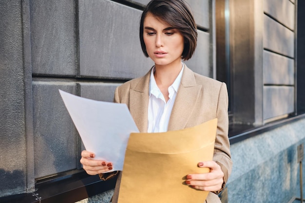 Free photo young serious businesswoman in suit thoughtfully working with papers on city street