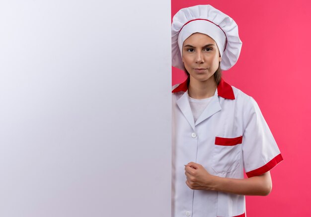 Young serious caucasian cook girl in chef uniform stands behind white wall on pink  with copy space