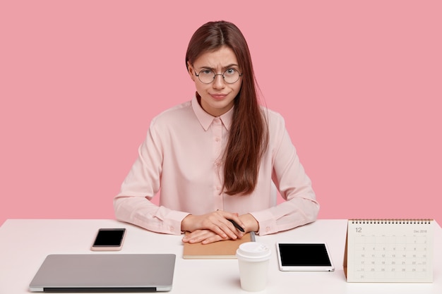 Young serious Caucasian woman wears spectacles, formal shirt, has everything on its place at table, surrounded with modern gadgets, notepad for records