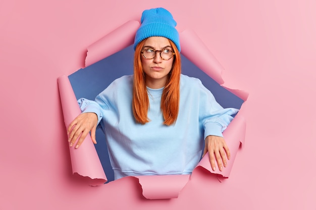 Free photo young serious skeptical redhead woman looks pensively aside has thoughtful expression thinks about something not very pleasant wears hat and sweater breaks through paper