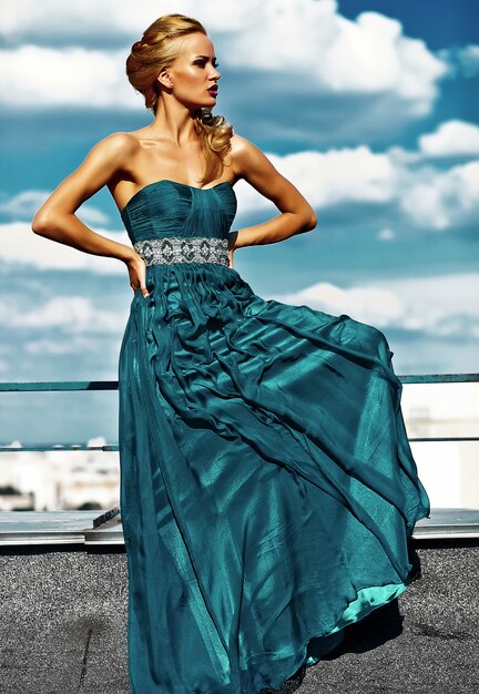 Young sexy blond woman model  in evening dress posing on blue sky background