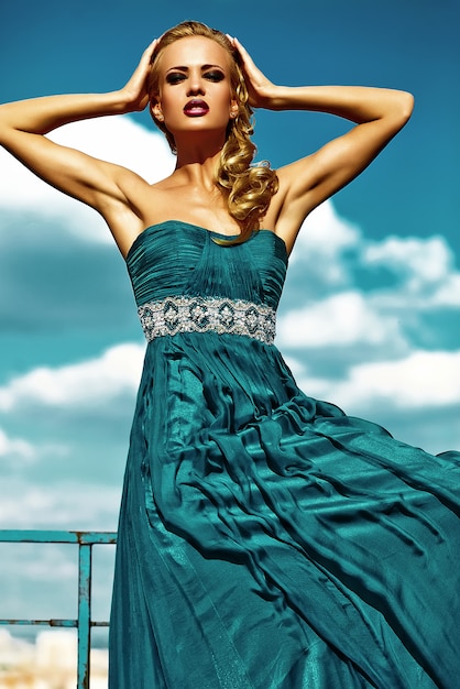 Free photo young sexy blond woman model  in evening dress posing on blue sky background