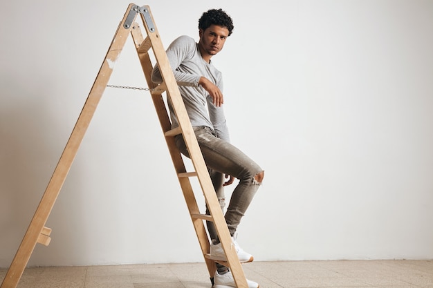 Free photo young sexy latin guy wears blank grey henley longsleeve and jeans sits on wooden ladder , isolated on white