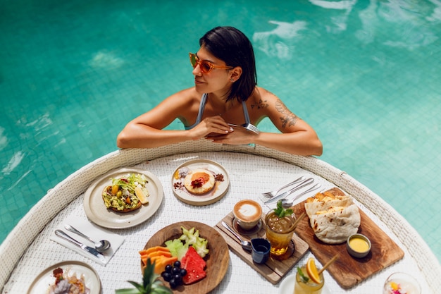 Free Photo young  sexy woman with tattoo  in a bathing suit having breakfast in a private pool.girl relaxing in the pool drinking coffee  and eating fruits. fruit plate, smoothie bowl by the hotel pool.