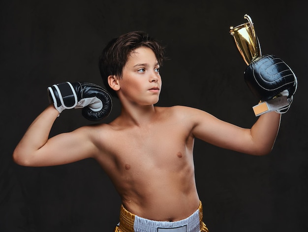 Young shirtless boxer champion wearing gloves holds a winner's cup showing muscles. Isolated on a dark background.