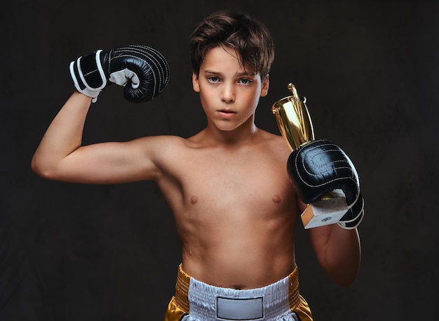 Young shirtless boxer champion wearing gloves holds a winner's cup showing muscles. Isolated on a dark background.