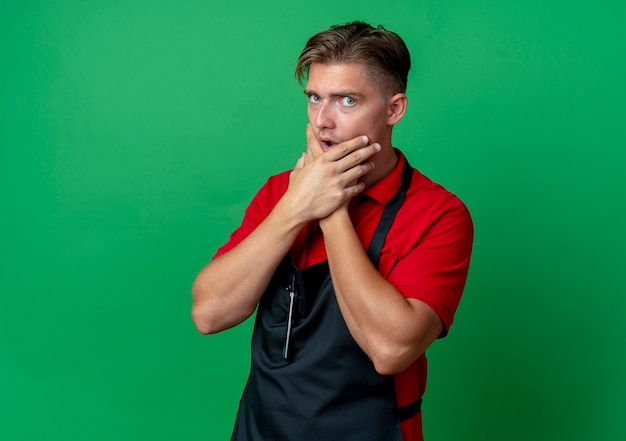 Young shocked blonde male barber in uniform holds chin with two hands isolated on green space with copy space