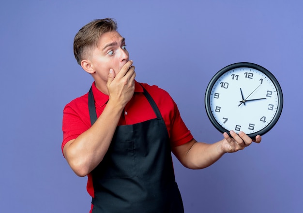 Young shocked blonde male barber in uniform holds and looks at clock isolated on violet space with copy space