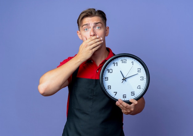 Young shocked blonde male barber in uniform puts hand on mouth holding clock isolated on violet space with copy space