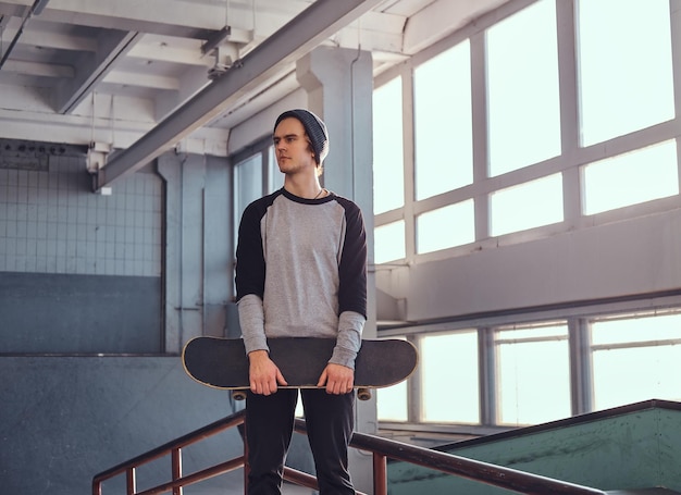 Young skateboarder standing next to a grind rail in skatepark indoors,  holding his board and looking away.