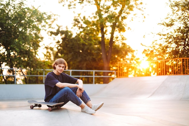 Free photo young skater in black tshirt and jeans dreamily looking in camera spending time with skateboard at modern skate park with sunset on background