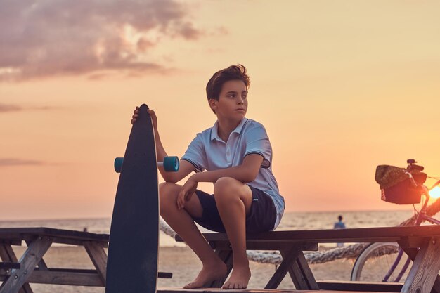 Young skater boy dressed in t-shirt and shorts sitting on a bench against the background of a seacoast at a bright sunset.