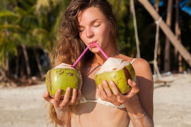 Young skinny woman in white bikini swimwear holding coconuts, smiling, sunbathing on tropical beach.