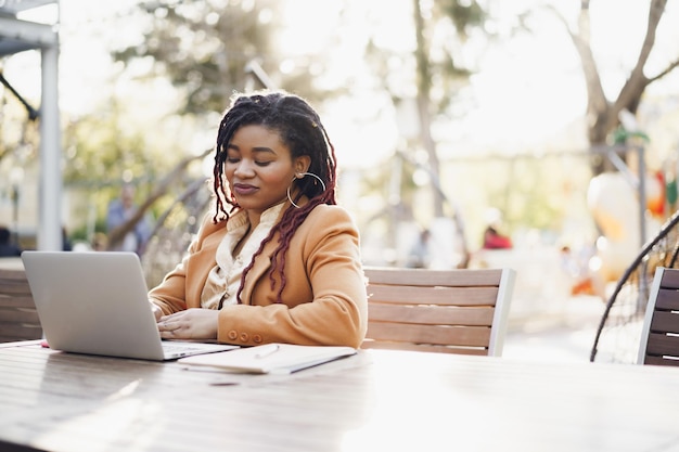 Young smiling african american woman sitting at the table in street cafe and using laptop