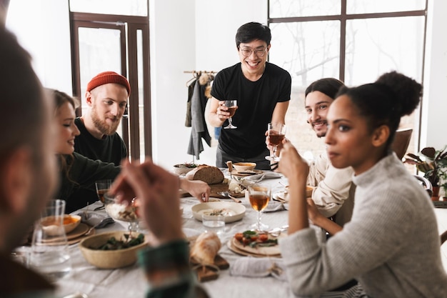 Young smiling asian man in eyeglasses and black Tshirt happily holding glass of wine in hand Group of attractive positive international friends spending time together on lunch in cozy cafe