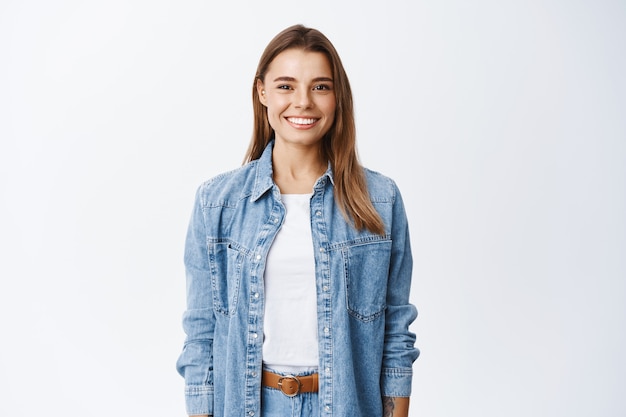 Free photo young smiling blond girl with perfect teeth staring at front, standing relaxed against white wall in casual clothes, lifestyle and emotions concept