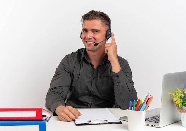 Young smiling blonde office worker man on headphones sits at desk with office tools using laptop puts finger on headphones isolated on white background with copy space