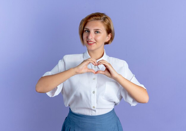 Young smiling blonde russian girl gestures heart hand sign isolated on purple background with copy space