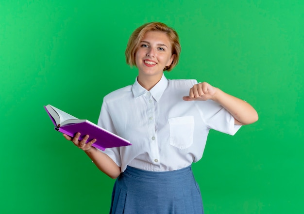 Free photo young smiling blonde russian girl holds book and points at herself isolated on green background with copy space