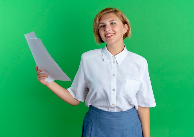 Free photo young smiling blonde russian girl holds paper sheets isolated on green background with copy space