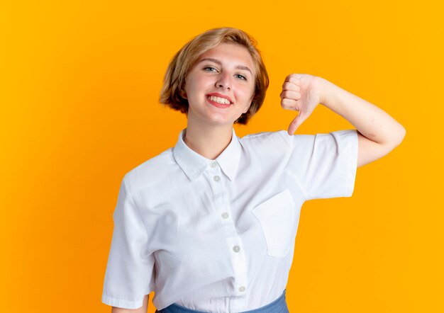 Young smiling blonde russian girl thumbs down isolated on orange background with copy space