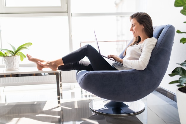 Young smiling brunette girl is sitting on modern chair near the window in light cozy room at home working on laptop in relaxing atmosphere