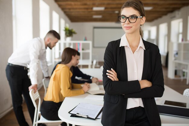 Young smiling business woman in eyeglasses and shirt dreamily looking aside with folded hands while spending time in office with colleagues on background