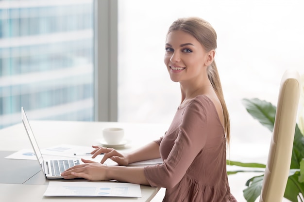 Free photo young smiling businesswoman sitting at office desk looking at camera