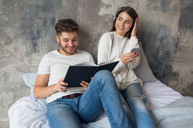 Young smiling couple sitting on bed at home in casual outfit reading book wearing jeans, man reading book, woman listening to music on headphones, spending romantic time together