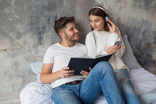 Young smiling couple sitting on bed at home in casual outfit reading book wearing jeans, man reading book, woman listening to music on headphones, spending romantic time together