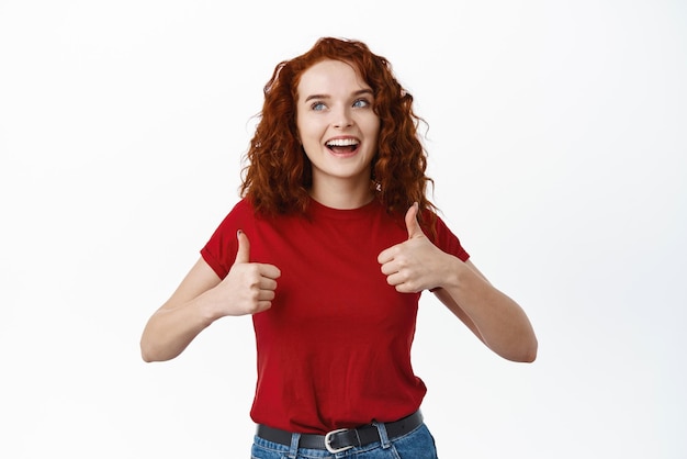 Free Photo young smiling curlyhaired ginger girl show thumbs up and look at upper left corner praise product promo recommending shop standing against white background